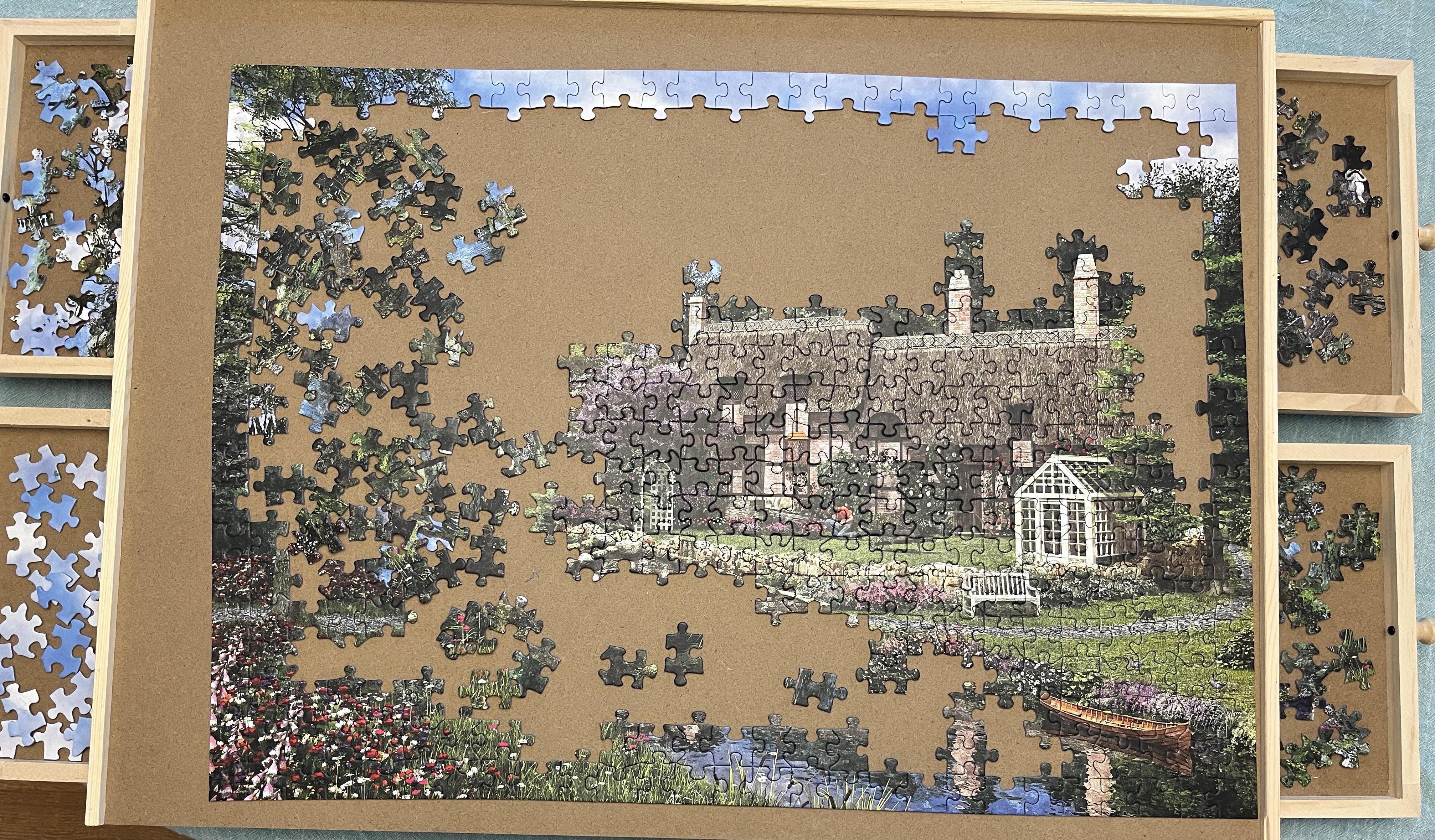A picture of a puzzle board with a partially completed puzzle. The edges and the cottage are completed but the rest is still scattered pieces